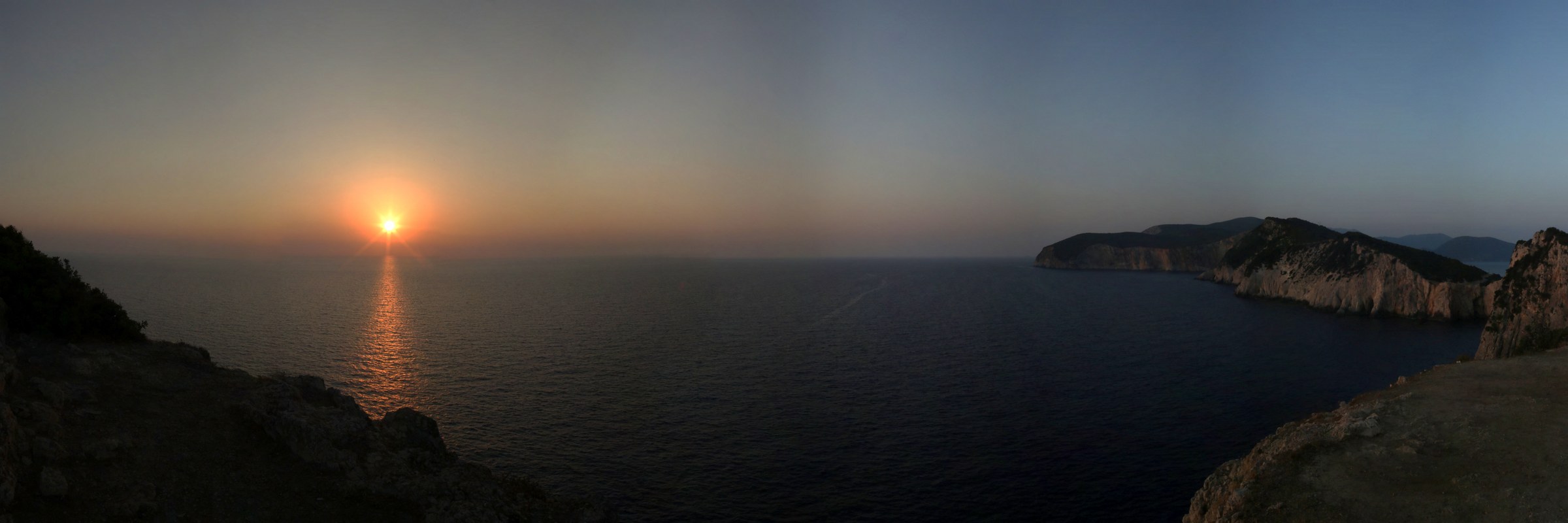 Il sole che tramonta sul mare, fra un capo di roccia e un'isola lontana